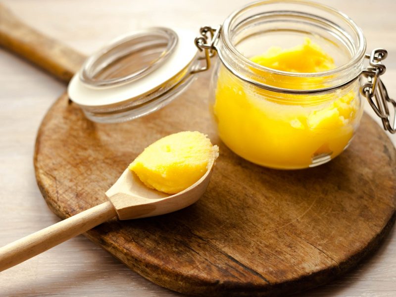 Ghee clarified butter desi yellow color in glass jar with spoon made from wood on natural wooden background close up view. Selective soft focus. Text copy space.